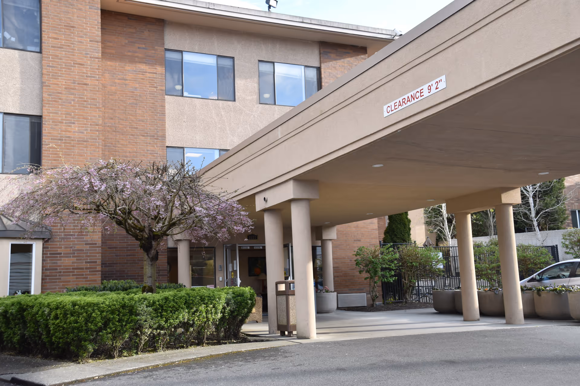 Exterior view of a multi-story brick and stucco building with a covered entrance supported by columns. There is a clearance sign indicating 9 feet 2 inches. A small tree with pink blossoms and green shrubs are visible near the entrance, along with a parked car to the right.