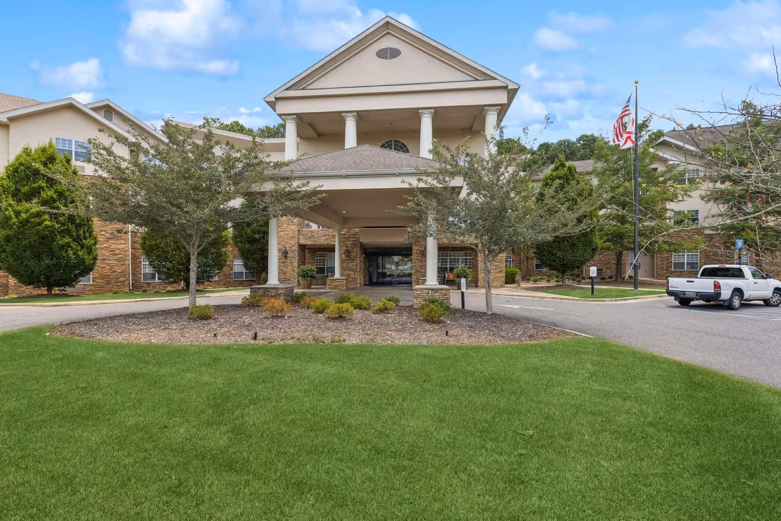 Front exterior view of a senior living facility with a covered entrance supported by white columns, surrounded by trees and landscaping, with a driveway and parked white pickup truck to the right. An American flag is visible on a flagpole near the entrance.