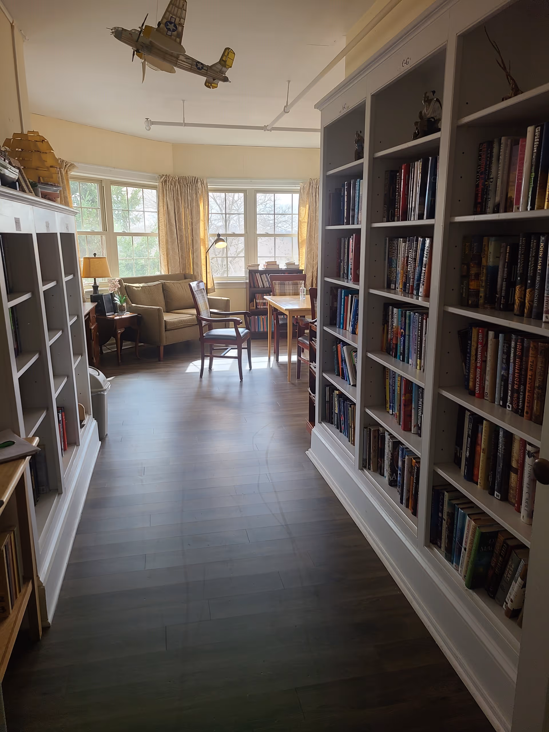 A cozy library room with bookshelves filled with books on the right and left sides. At the end of the room, there is a sitting area with a beige couch, two wooden chairs, a small table, and a lamp near large windows with yellow curtains. A model airplane hangs from the ceiling.