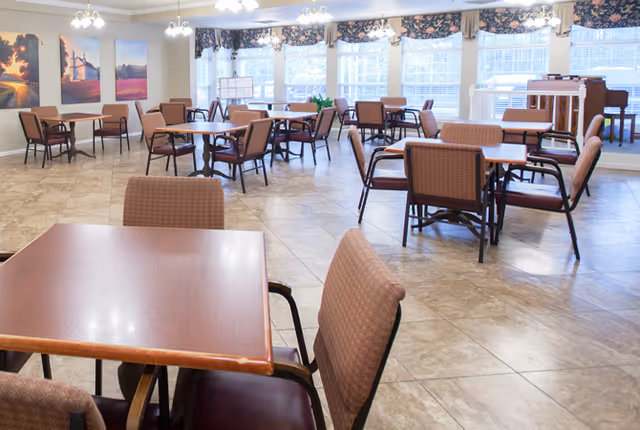 A spacious dining room with multiple square wooden tables and cushioned chairs arranged neatly. Large windows with floral valances allow natural light to fill the room. The floor is tiled, and there are paintings on the wall and a piano in the background.