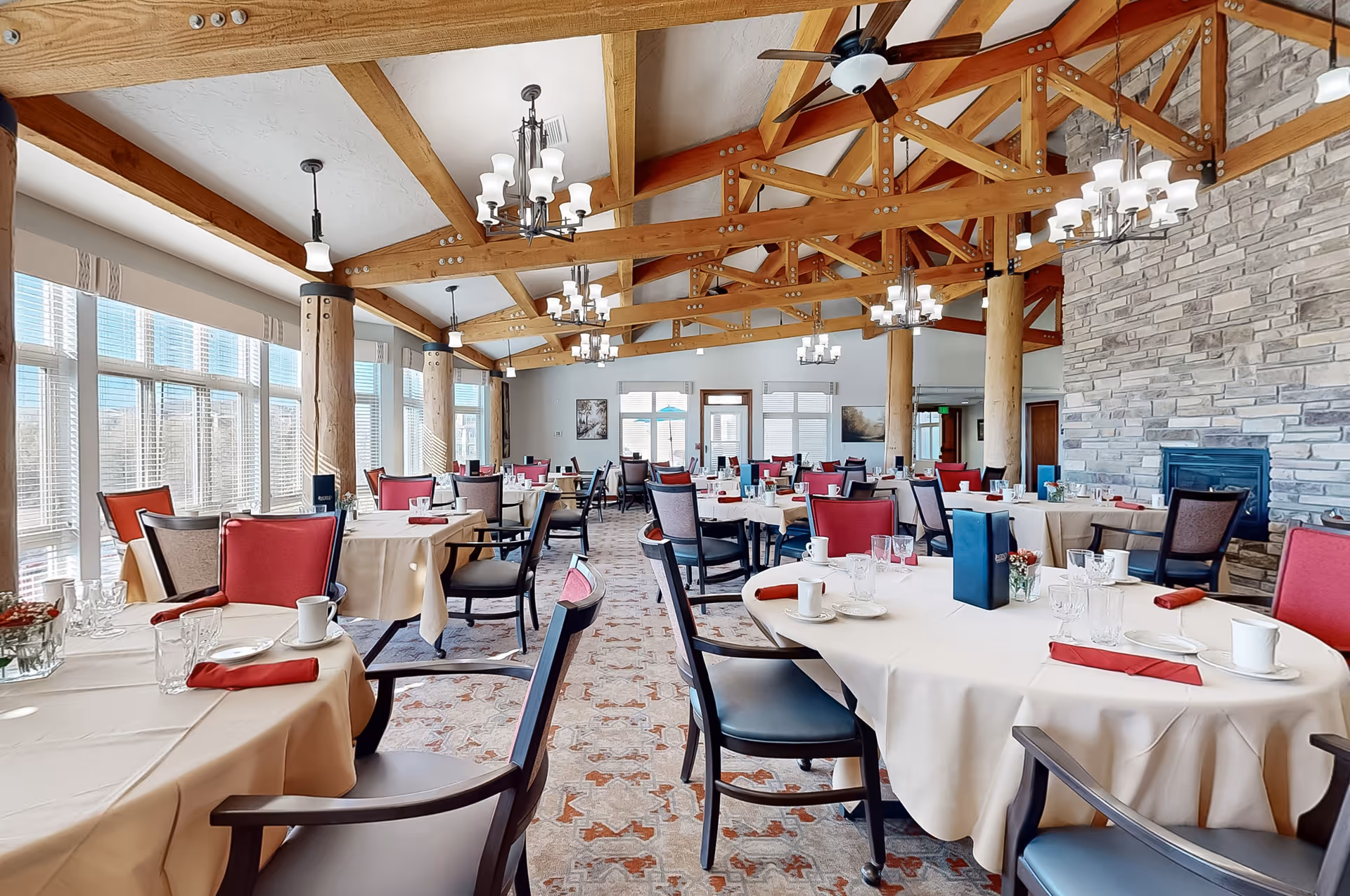 A spacious dining room with multiple round and rectangular tables covered with beige tablecloths, set with white plates, cups, glasses, and red napkins. The room features large windows with blinds, wooden beams on the ceiling, chandeliers, ceiling fans, and a stone fireplace on the right wall. Chairs have a mix of red and gray upholstery.