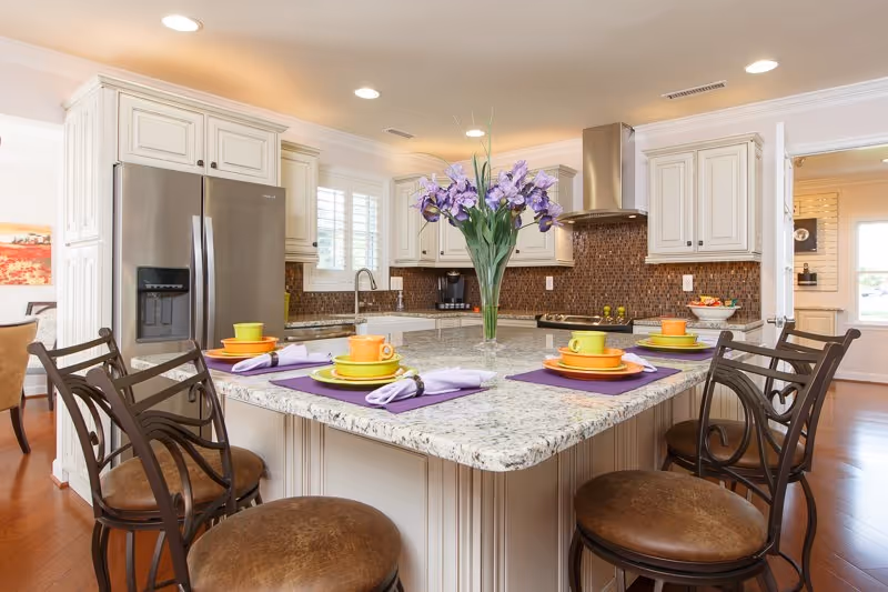 Spacious traditional kitchen featuring a granite island with bar stools, colorful place settings, and a vase of purple flowers.
