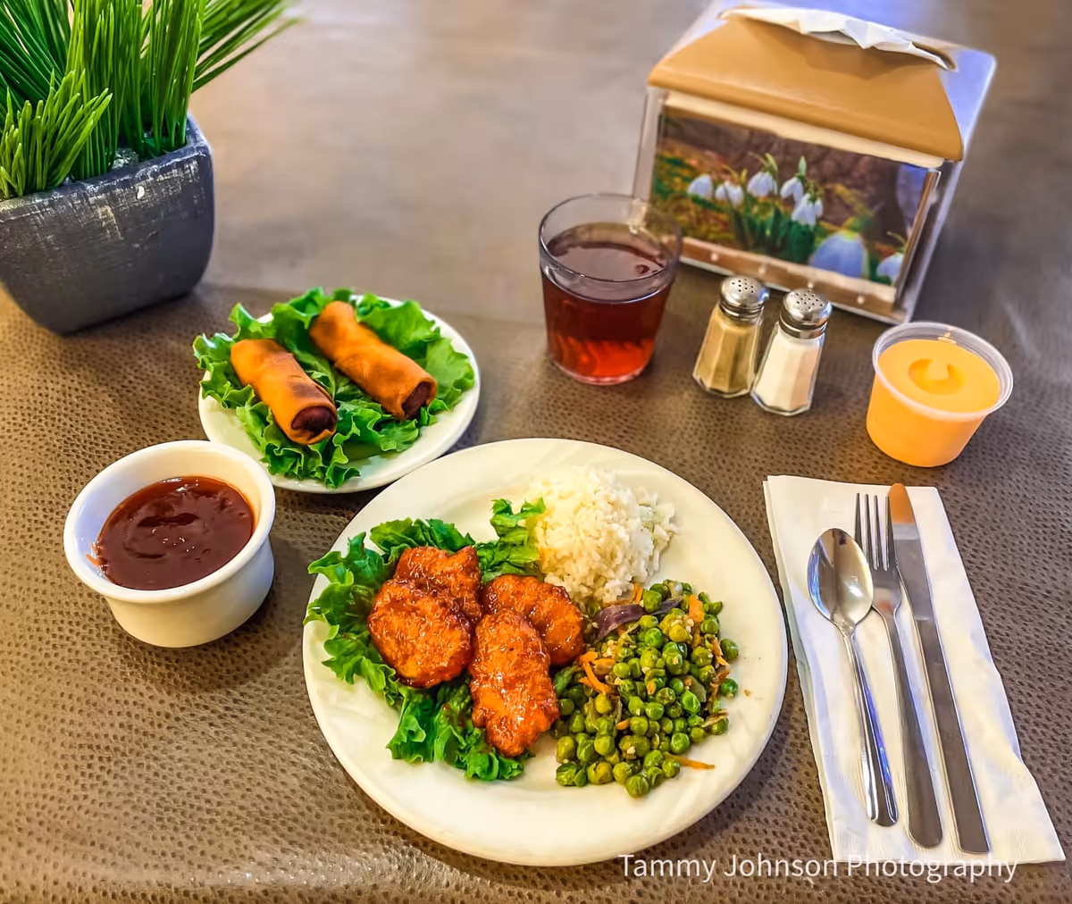 A plated meal of rice, peas and fried chicken with spring rolls, dipping sauce, drink and utensils on a dining table.