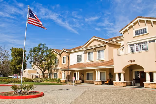 Exterior view of a two-story senior living facility building with beige and light brown walls, a tiled roof, and several windows. An American flag on a flagpole is prominently displayed in the front near a landscaped area with bushes and trees. The sky is clear with a few clouds.