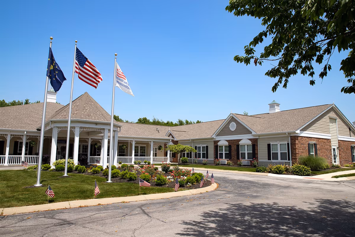 Exterior view of The Commons at Honey Creek senior living facility showing a single-story building with a covered entrance, three flagpoles with flags, a landscaped garden with small American flags, and a clear blue sky.