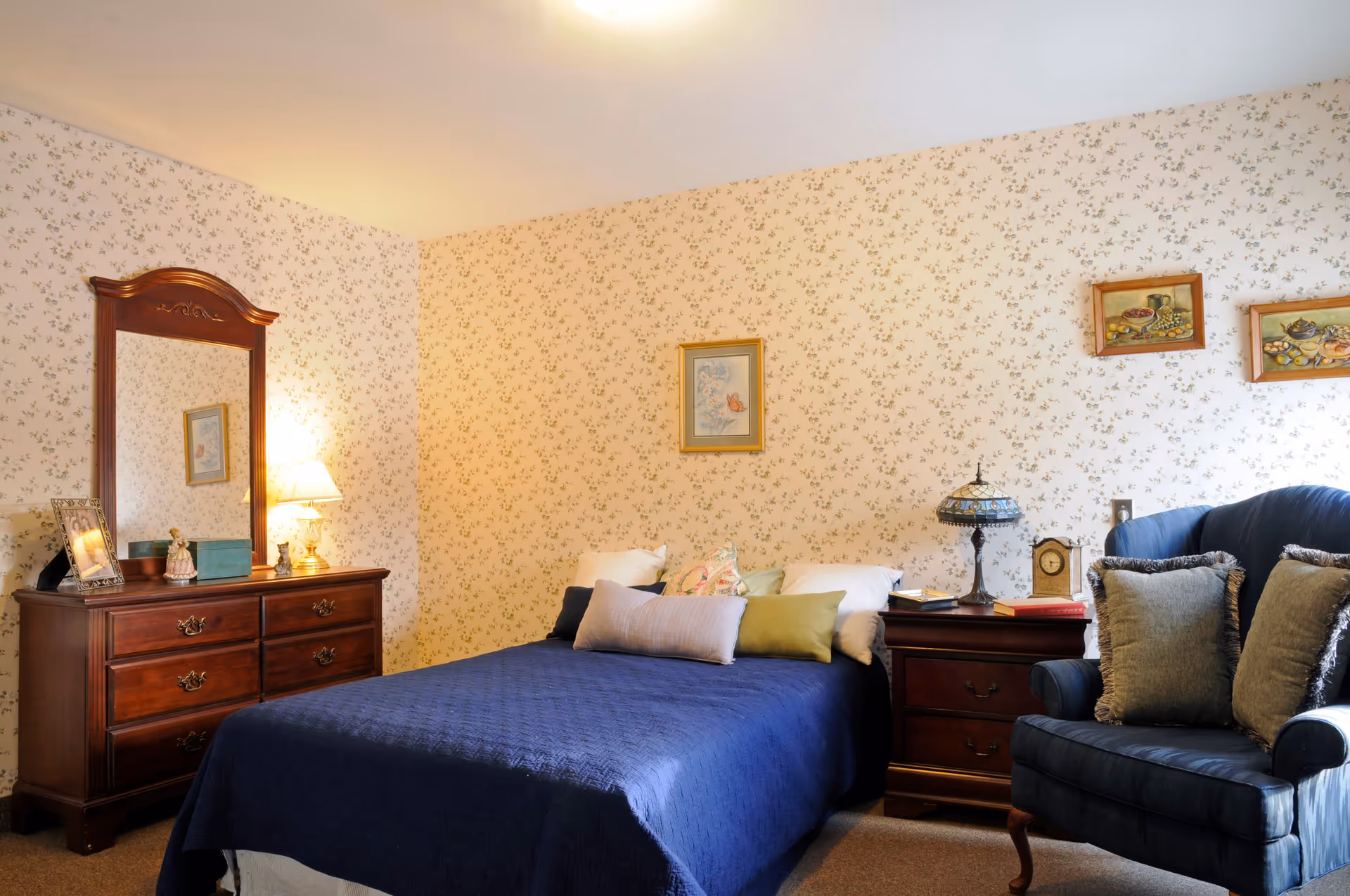 A cozy bedroom with floral wallpaper, featuring a bed with a dark blue quilt and multiple pillows. To the left is a wooden dresser with a large mirror and a table lamp. To the right of the bed is a wooden nightstand with a decorative lamp, clock, and books. A blue upholstered armchair with two pillows is positioned next to the nightstand. Several framed pictures hang on the walls.