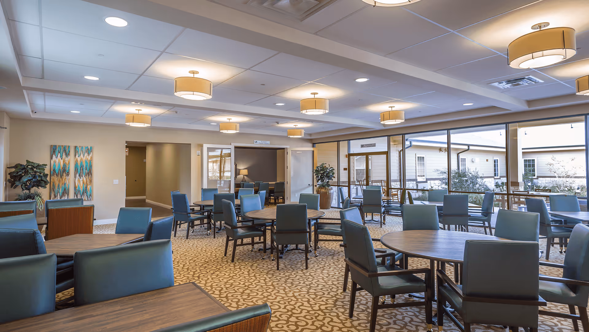 A spacious dining room with multiple round and rectangular tables surrounded by teal upholstered chairs. The room has large windows letting in natural light, beige walls, patterned carpet, and ceiling lights with drum shades. There are decorative plants and artwork on the walls.