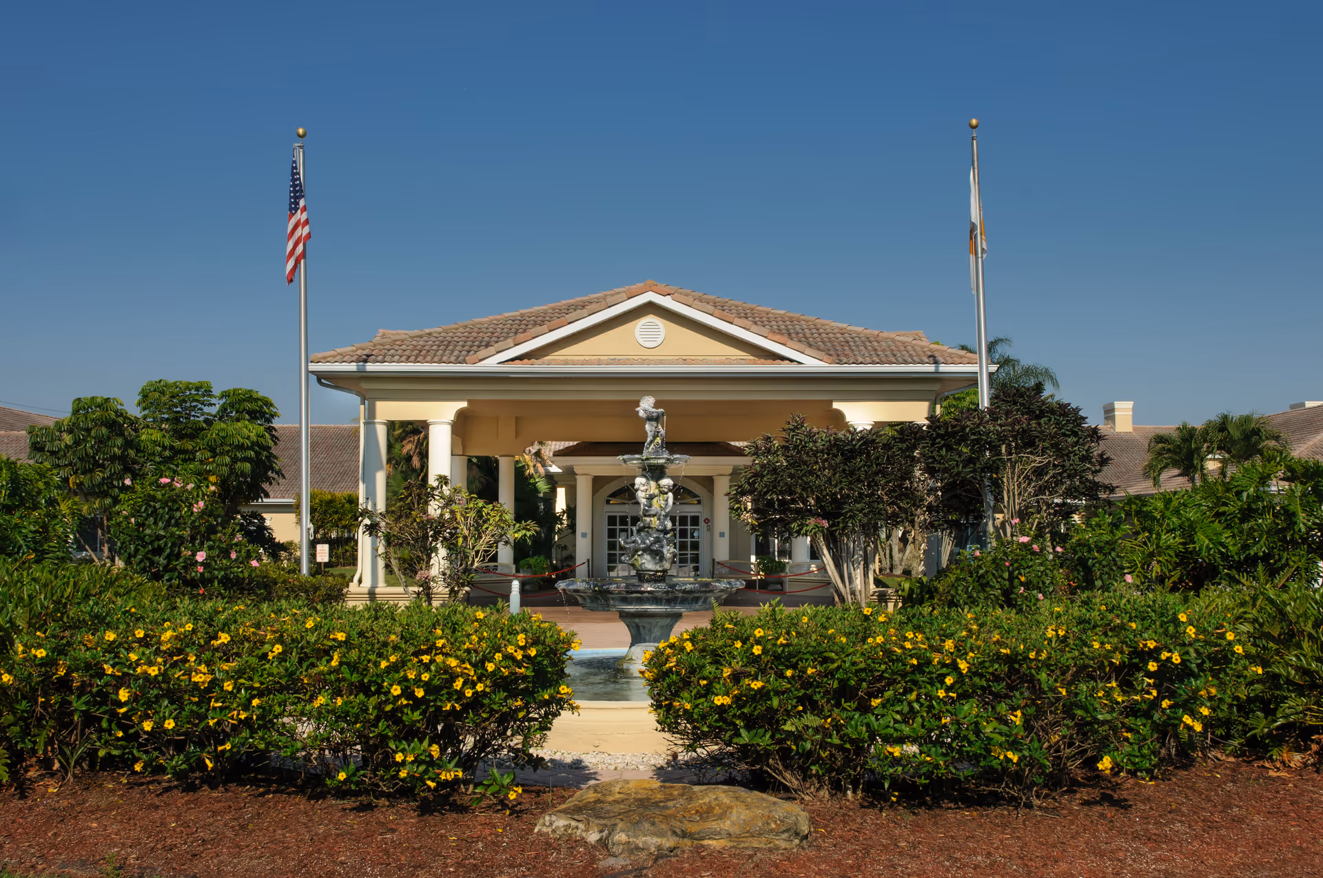 Front entrance of a beige building with columns, a central fountain, two flagpoles, and landscaped flowerbeds under a clear blue sky.