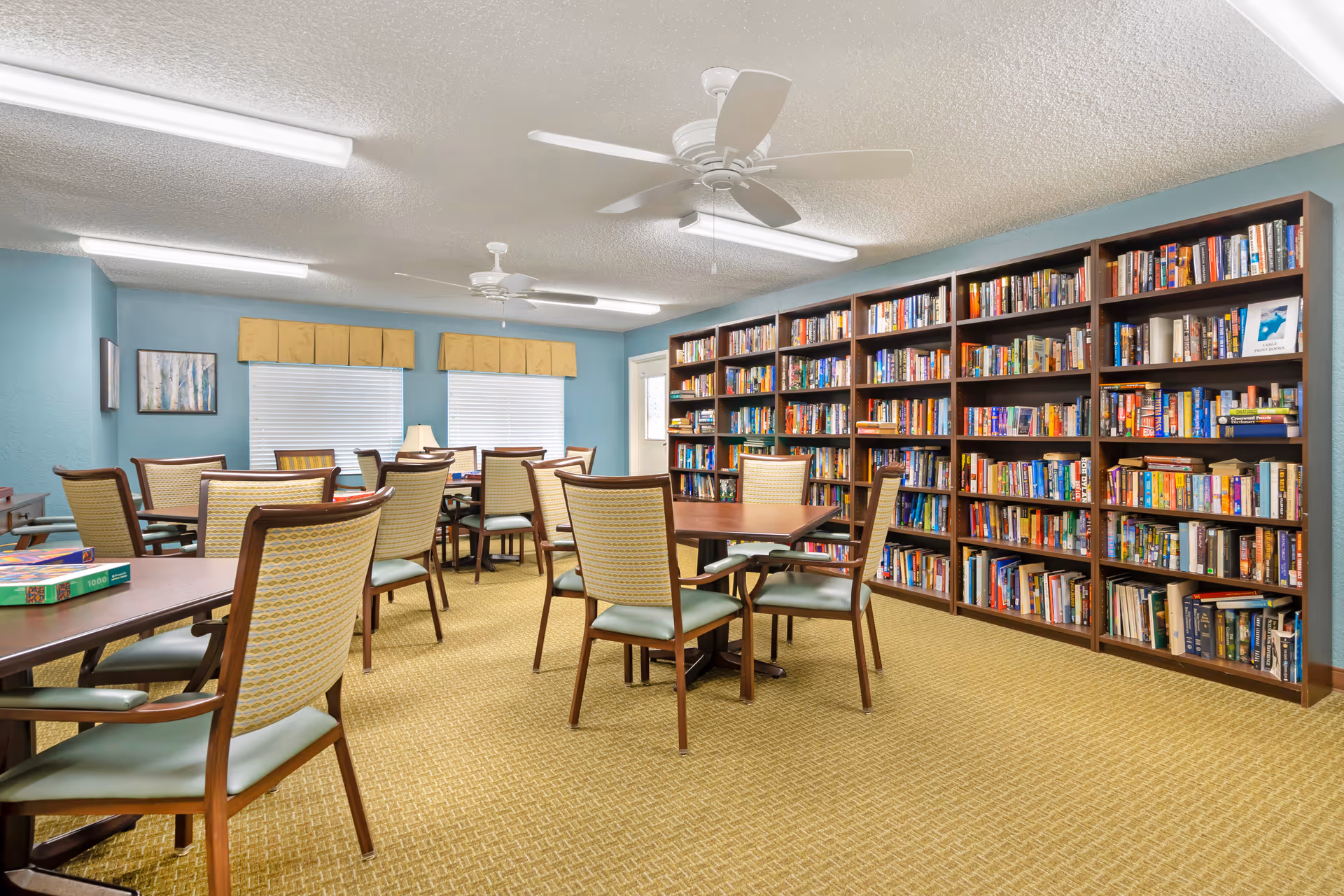A bright room with light blue walls and beige carpet featuring multiple wooden tables and chairs with green cushions. One wall is lined with large bookshelves filled with books. The room has two ceiling fans and fluorescent lighting, with two windows covered by yellow valances.