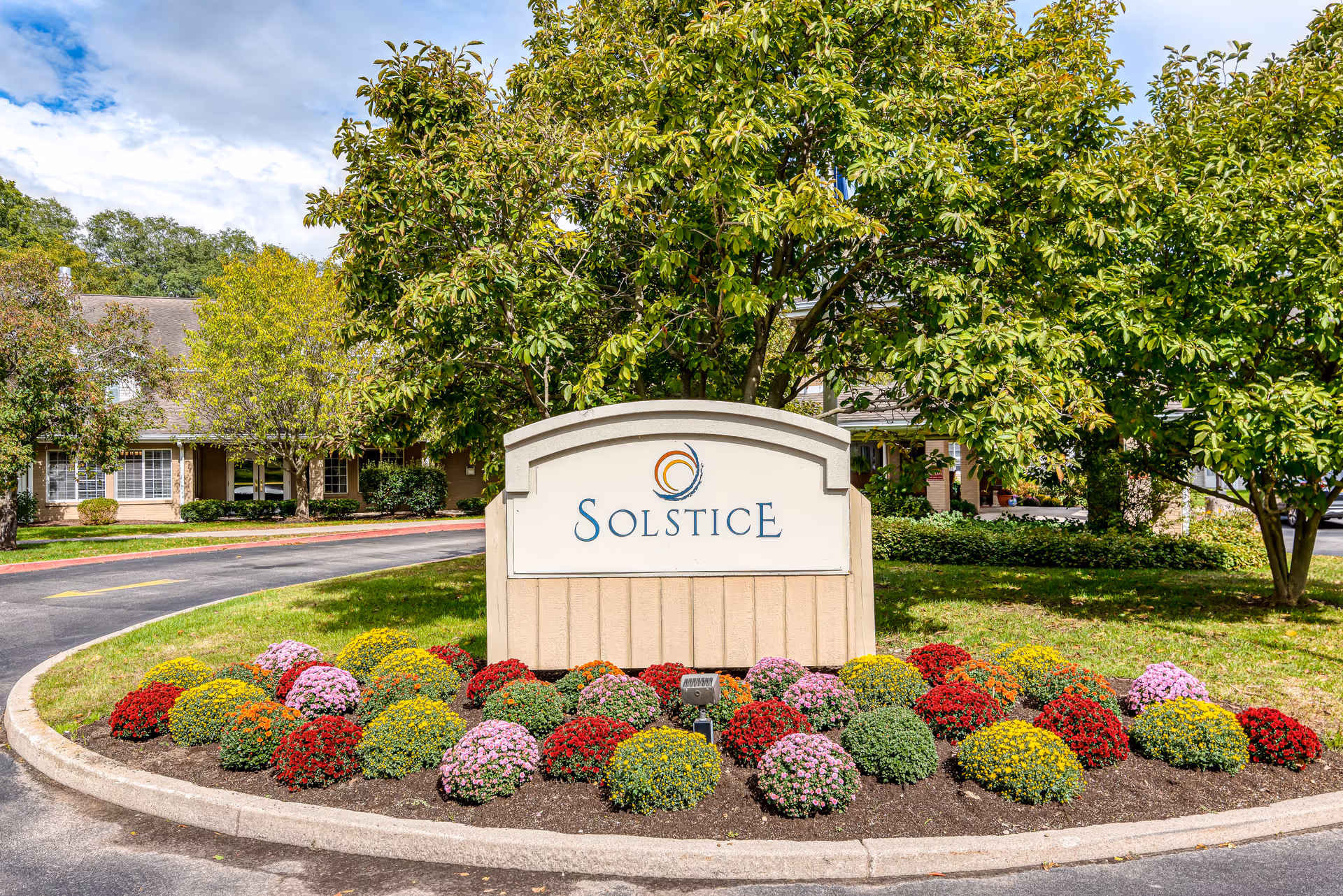 Entrance sign reading 'Solstice' set in a landscaped island of multicolored flowers with trees and the facility building behind it.