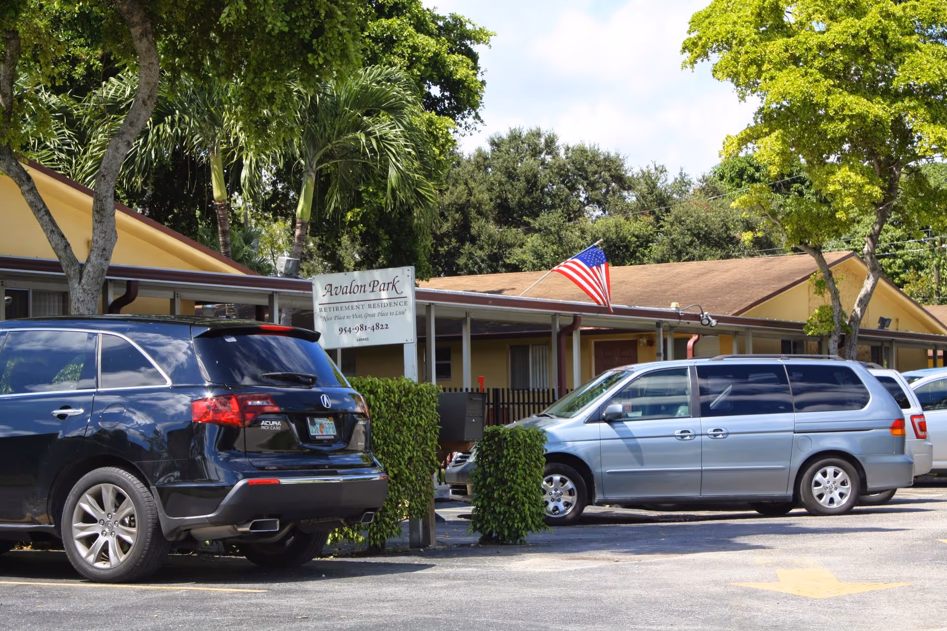 Parking lot in front of Avalon Park Retirement Residence with several parked vehicles, including a black Acura SUV and a silver minivan. The building is a single-story structure with a beige exterior and an American flag displayed on the roof. Trees and greenery surround the area under a partly cloudy sky.