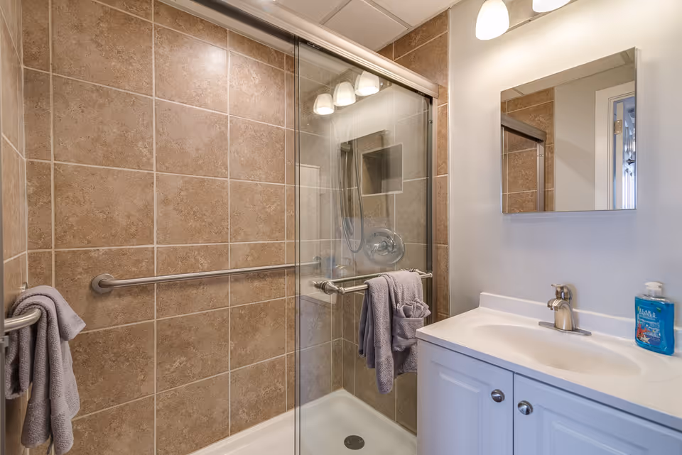 A bathroom with a tiled walk-in shower featuring a glass sliding door and a metal grab bar. There are two gray towels hanging on the grab bars. Next to the shower is a white vanity with a sink, a silver faucet, a mirror above it, and a bottle of blue liquid soap on the countertop. The walls are white and the lighting fixture above the mirror has three lights.