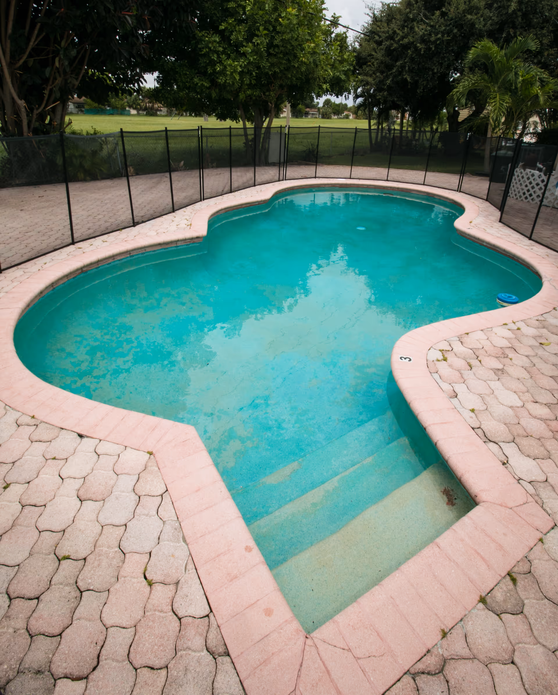 A small outdoor swimming pool with light blue water, surrounded by a pinkish brick border and a paved patio area. The pool has steps leading into the water and is enclosed by a black safety fence. Trees and green grass are visible in the background.