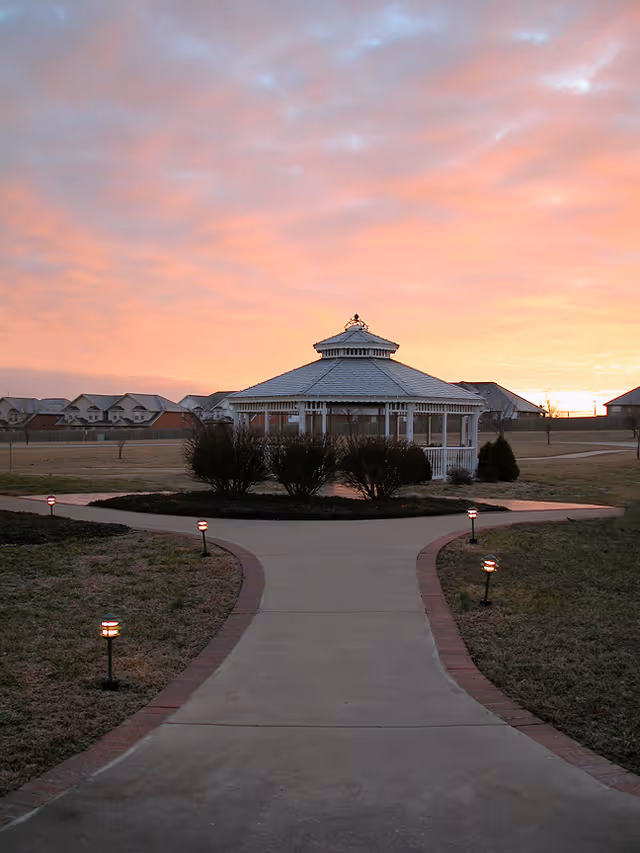 A paved walkway leading to a white gazebo surrounded by bushes and small lights on either side, with a sunset sky in the background and houses visible in the distance.