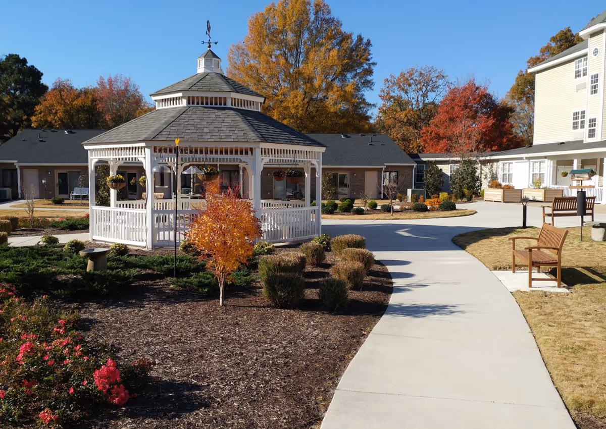 Outdoor area of Mennowood Retirement Community featuring a white gazebo with a weather vane on top, surrounded by landscaped gardens with bushes, small trees with autumn foliage, and a curved concrete pathway. There are wooden benches along the pathway and residential buildings in the background under a clear blue sky.
