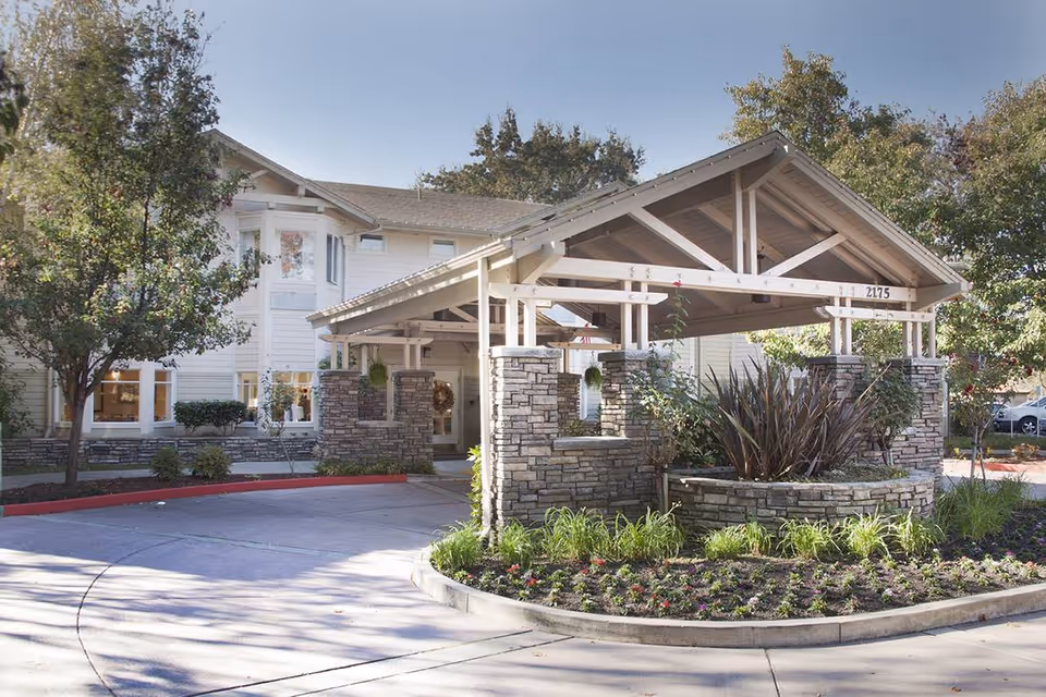 Exterior view of Ivy Park at Walnut Creek showing the entrance with a covered driveway supported by stone pillars, surrounded by landscaped greenery and trees under a clear sky.