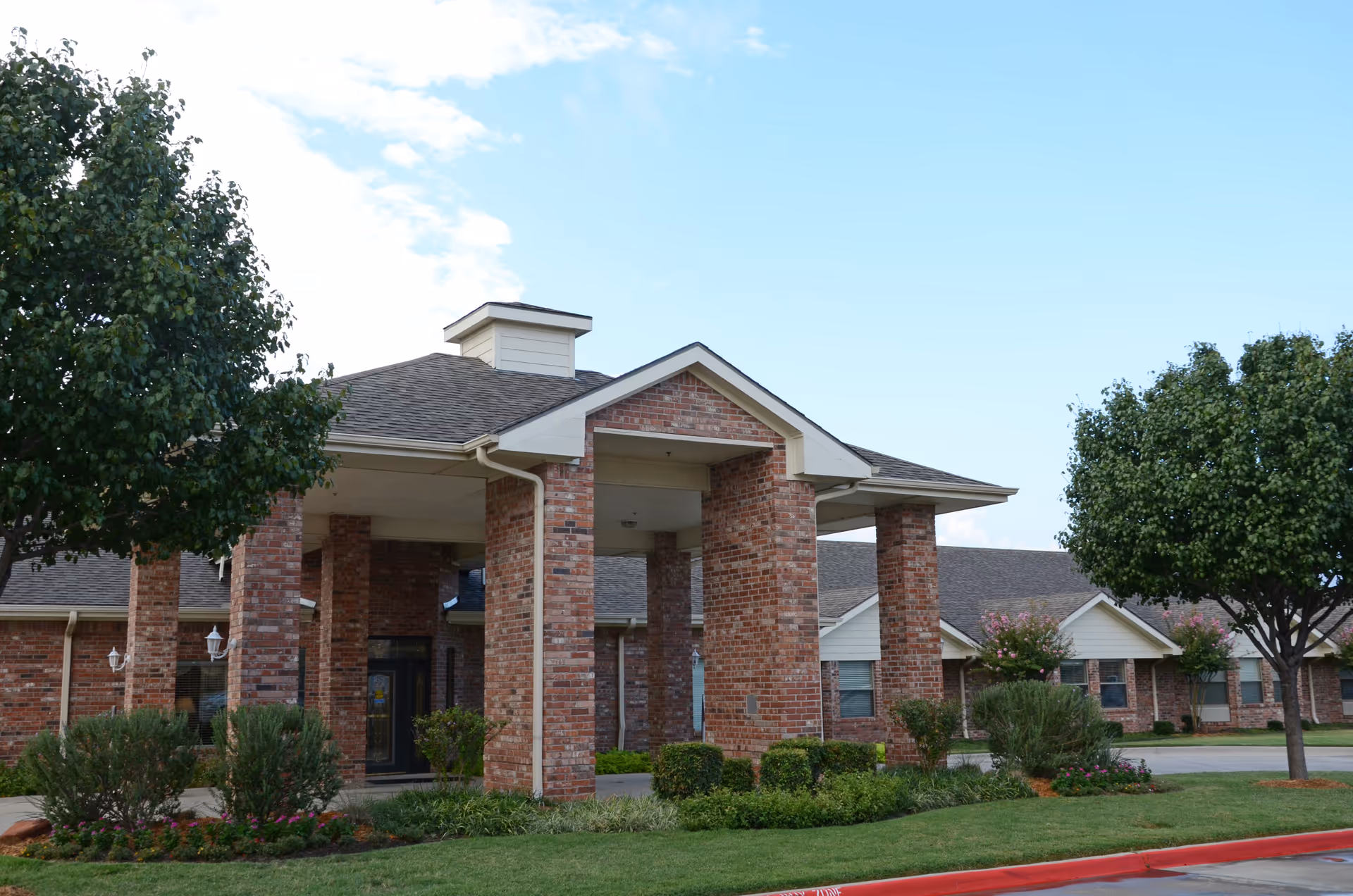 Brick-front nursing facility entrance with a covered portico, columns, landscaped lawn and trees under a blue sky.