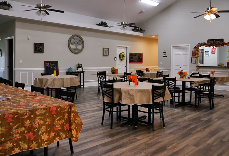 A bright assisted-living dining room with round tables covered in beige cloths, black chairs, ceiling fans, and autumn-themed centerpieces.