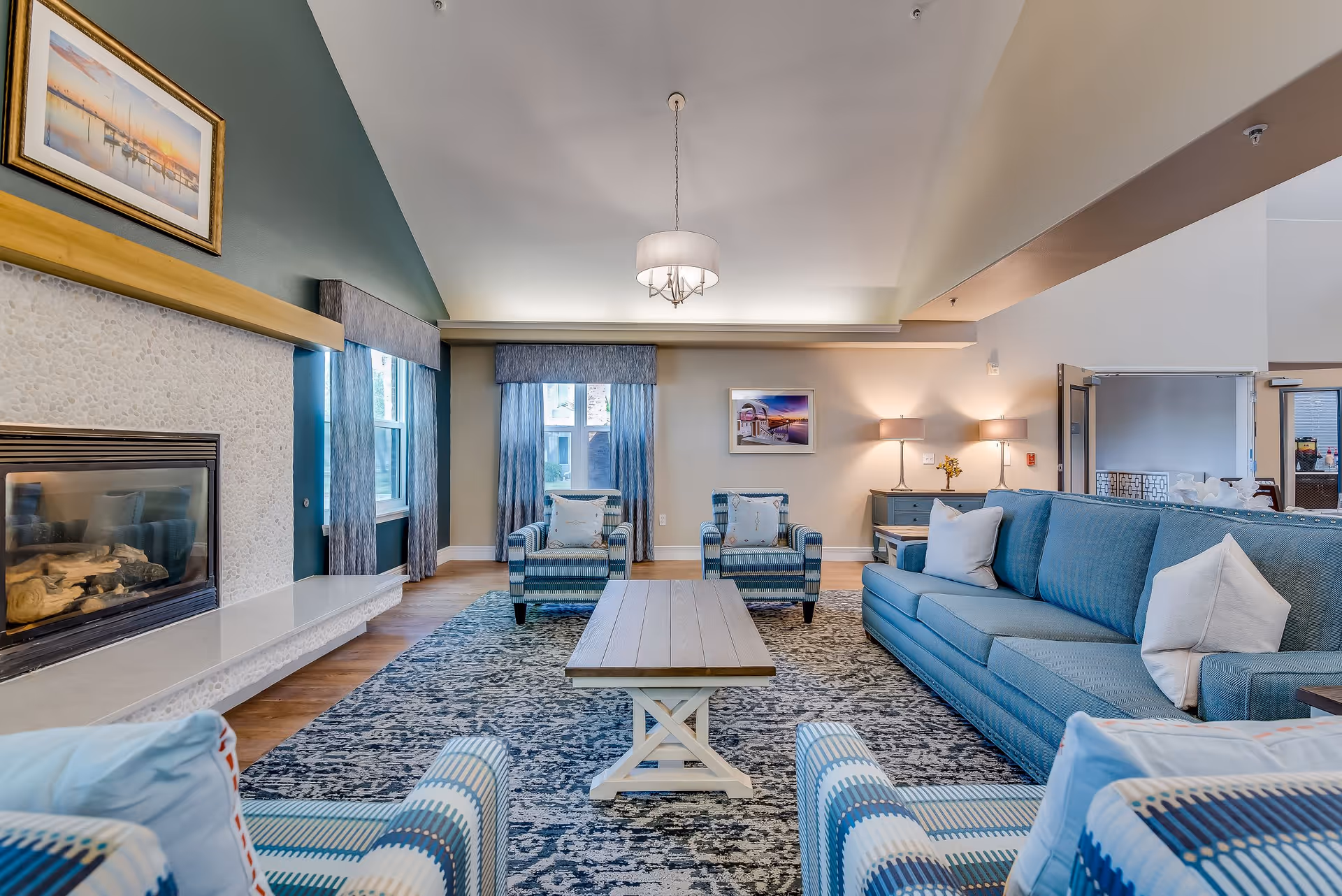 A spacious and well-lit living room with a high ceiling, featuring a blue sofa, two blue patterned armchairs, a wooden coffee table, a fireplace on the left wall, and two windows with blue curtains. The room has a large patterned rug, a chandelier hanging from the ceiling, and two table lamps on a sideboard against the far wall.