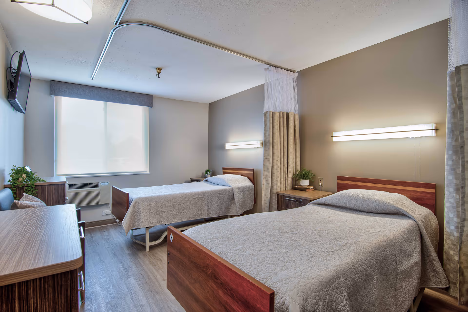 A clean and well-lit bedroom in a senior living facility with two single beds covered in beige quilts. Each bed has a wooden headboard and is accompanied by a nightstand with a small plant and lamp. A curtain divider hangs from the ceiling between the beds. There is a window with a white blind, a wall-mounted TV, an air conditioning unit, and a wooden dresser with a plant on top. The walls are painted beige and the floor has wood-like vinyl flooring.