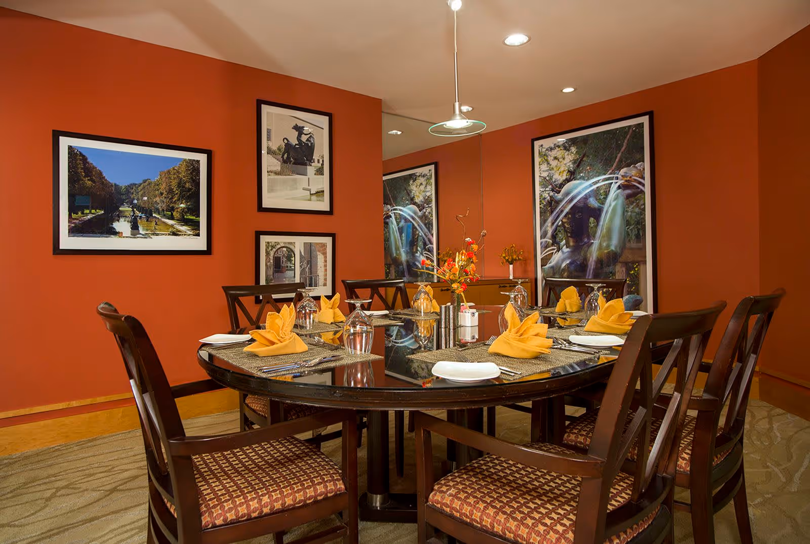 A dining room with a round glass-top table set for six people. The table has yellow folded napkins, glasses, plates, and silverware. The walls are painted orange and decorated with framed photographs. There is a modern ceiling light fixture above the table.