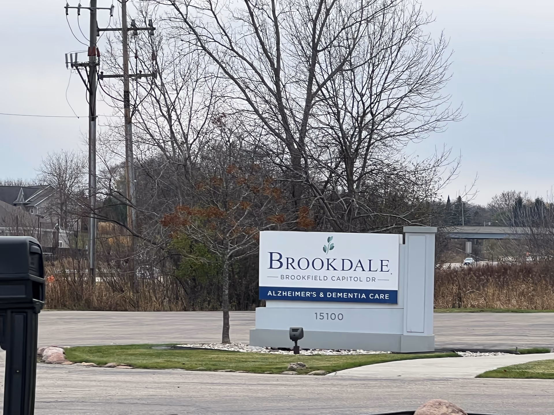 Outdoor view of a large white sign for Brookdale Brookfield Capitol Dr, an Alzheimer's and Dementia Care facility, located on a grassy area near a parking lot with leafless trees and power lines in the background.