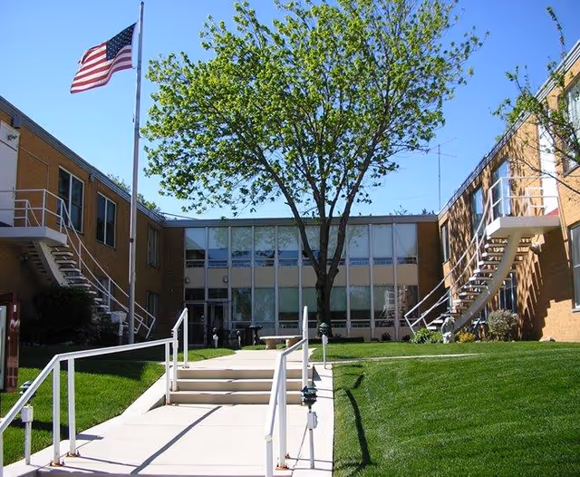 Outdoor courtyard area of King’s Gardens Campus of Perry Lutheran Homes with a large tree in the center, surrounded by a two-story brick building with exterior staircases. An American flag is flying on a flagpole to the left, and there is a paved walkway with handrails leading into the courtyard.