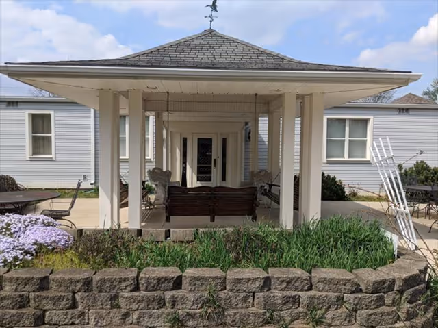 Outdoor covered seating area with a roof supported by white columns, featuring a wooden bench and two chairs. The area is surrounded by a low stone wall with green plants and purple flowers in front. The background shows a light gray building with windows and a door under the covered area.