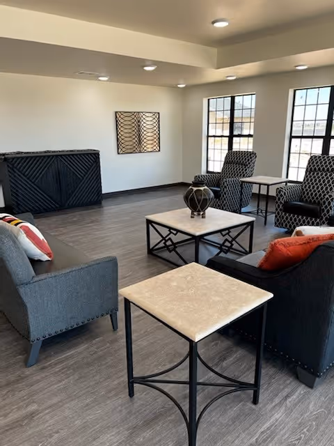 A modern living room area with gray and black armchairs and sofas arranged around two square coffee tables with light-colored tops. The room has large windows letting in natural light, a decorative wall piece, and a dark wooden cabinet against one wall. The flooring is a wood-like material and the ceiling has recessed lighting.