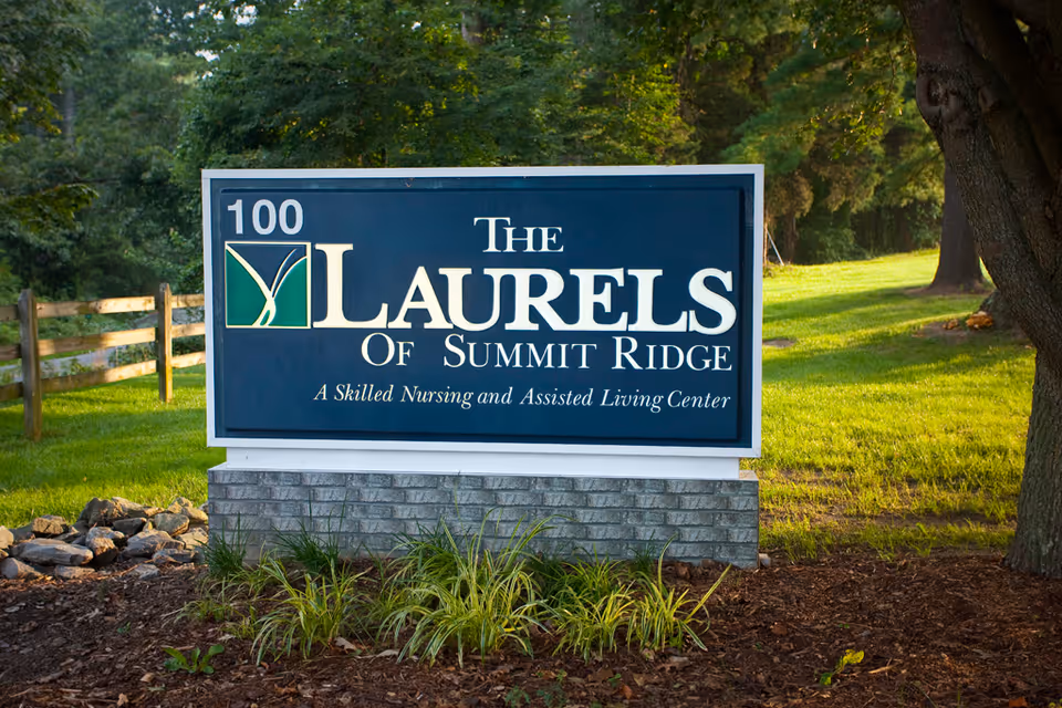 A large outdoor sign for The Laurels of Summit Ridge, a skilled nursing and assisted living center, situated on a grassy area with trees and a wooden fence in the background.