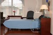 A hospital-style bed with a blue bedspread in a room with wooden flooring. Next to the bed is a wooden nightstand with a lamp and a telephone. Behind the bed is a high-backed chair and a small table with a vase of flowers. Large windows with vertical blinds allow natural light into the room.