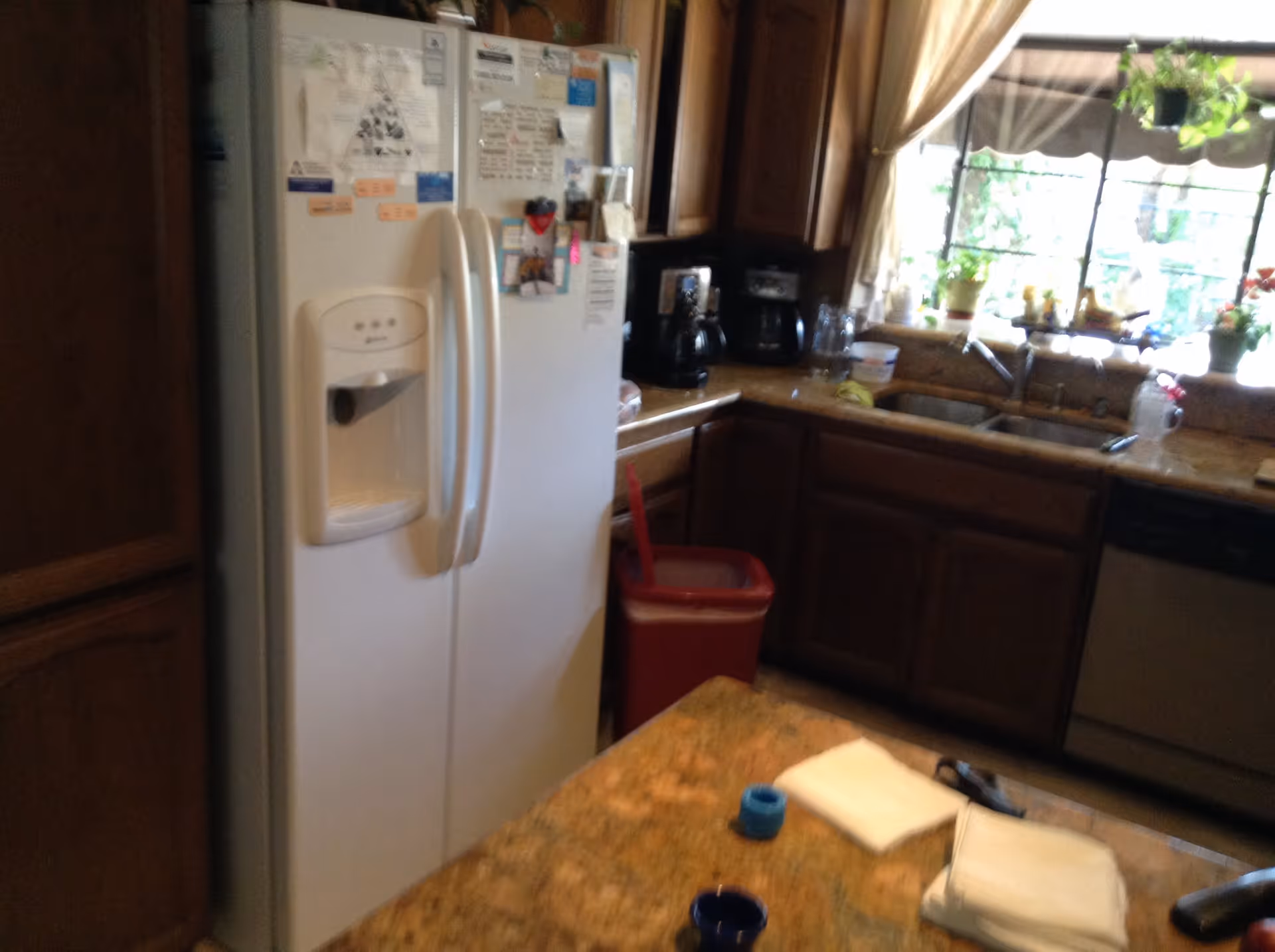 A kitchen with a white refrigerator covered in magnets and notes, wooden cabinets, a countertop with a coffee maker and other items, a double sink under a window with plants on the windowsill, and a kitchen island with folded napkins and a cup.