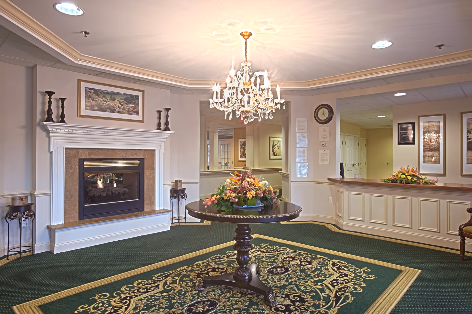 Elegant senior living lobby with a chandelier, central round table holding a floral arrangement, fireplace, and reception desk.