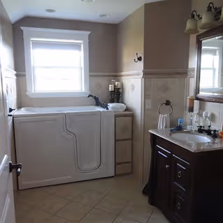 A bathroom with a walk-in bathtub on the left side, a window above the tub, and a dark wooden vanity with a sink and mirror on the right side. The walls are tiled halfway up with decorative tiles, and the floor is tiled with light brown tiles.