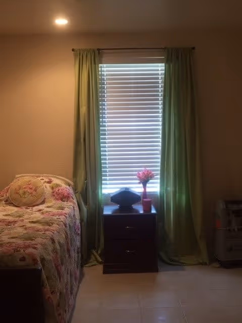 Small bedroom with a single floral-covered bed, a nightstand beneath a window with green curtains and a pink potted flower.