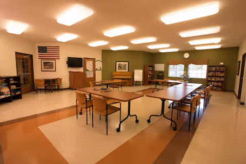 A well-lit community room with several tables arranged in a U-shape, surrounded by chairs. The room features a piano, bookshelves, an American flag on the wall, a television, and a whiteboard. Large windows with valances allow natural light to enter the space.