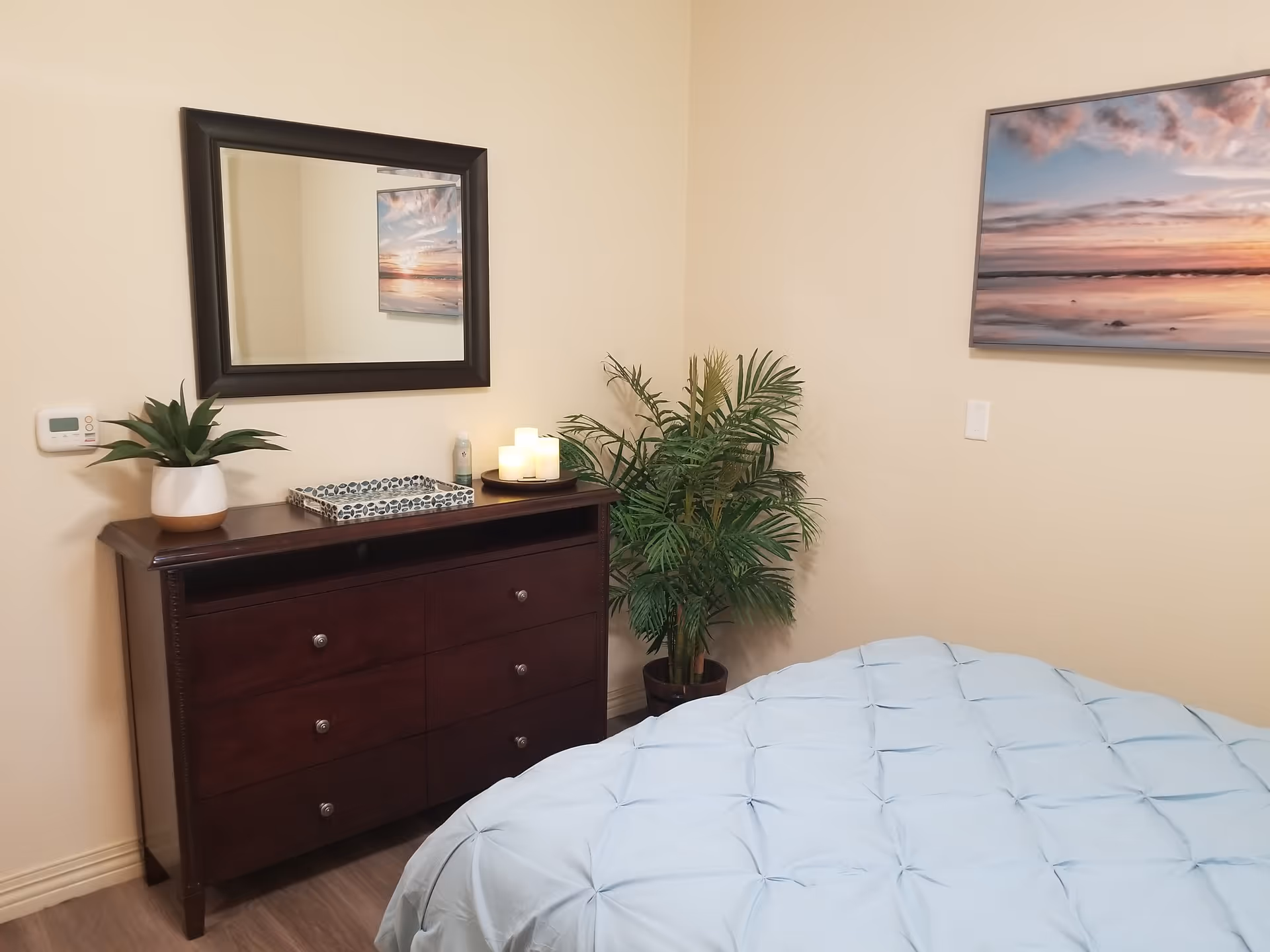 A bedroom corner featuring a dark wooden dresser with six drawers, a rectangular mirror above it, a potted plant, a decorative tray, and three lit candles. Next to the dresser is a large green potted plant. Part of a bed with a light blue comforter is visible in the foreground. A framed picture of a sunset over water hangs on the beige wall.