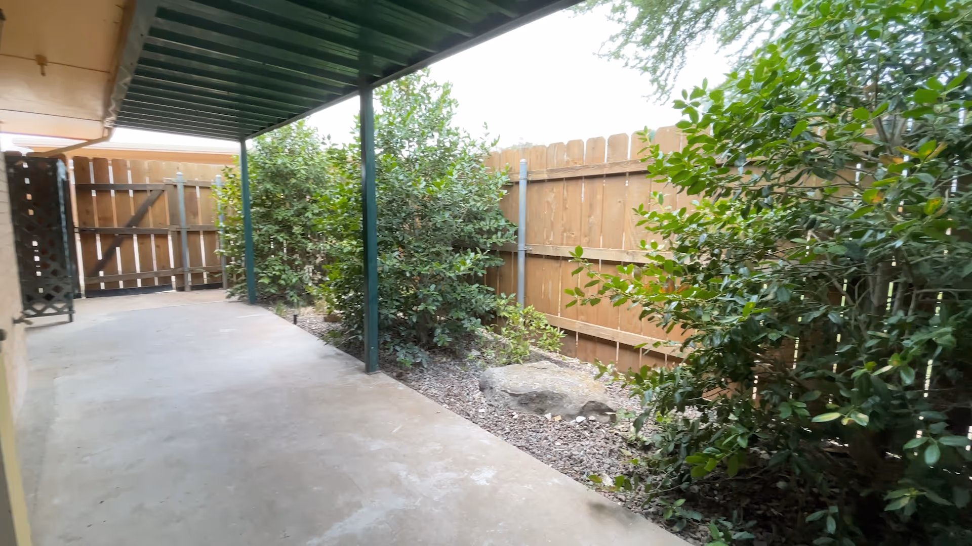 Covered concrete patio/walkway beside a wooden fence with shrubs and a gate.