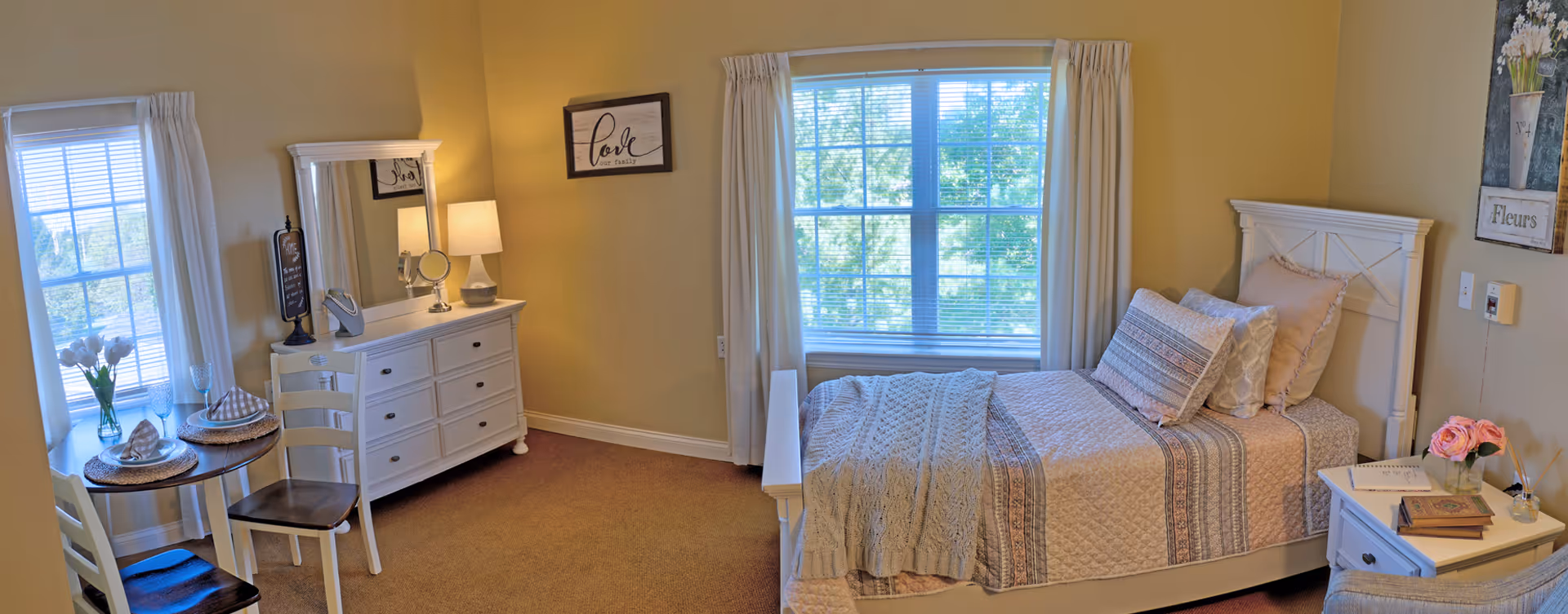 A cozy bedroom in a senior living facility with a single bed covered in a quilt and pillows, a white dresser with a mirror and a lamp, a small round dining table set for two near a window, and soft yellow walls decorated with framed artwork.