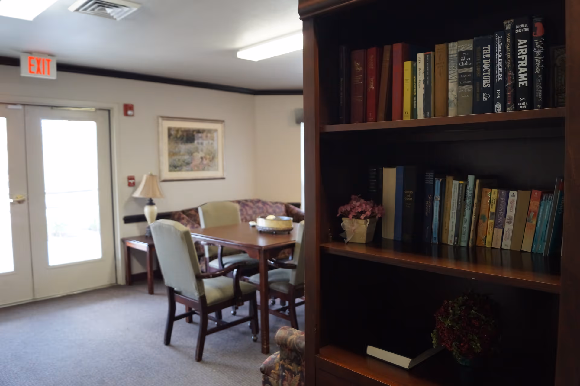 Interior view of a common area in a senior living facility featuring a wooden bookshelf filled with books and decorative plants on the right side. In the background, there is a wooden table with four upholstered chairs around it, a lamp on a side table, a framed picture on the wall, and double glass doors with an exit sign above them.