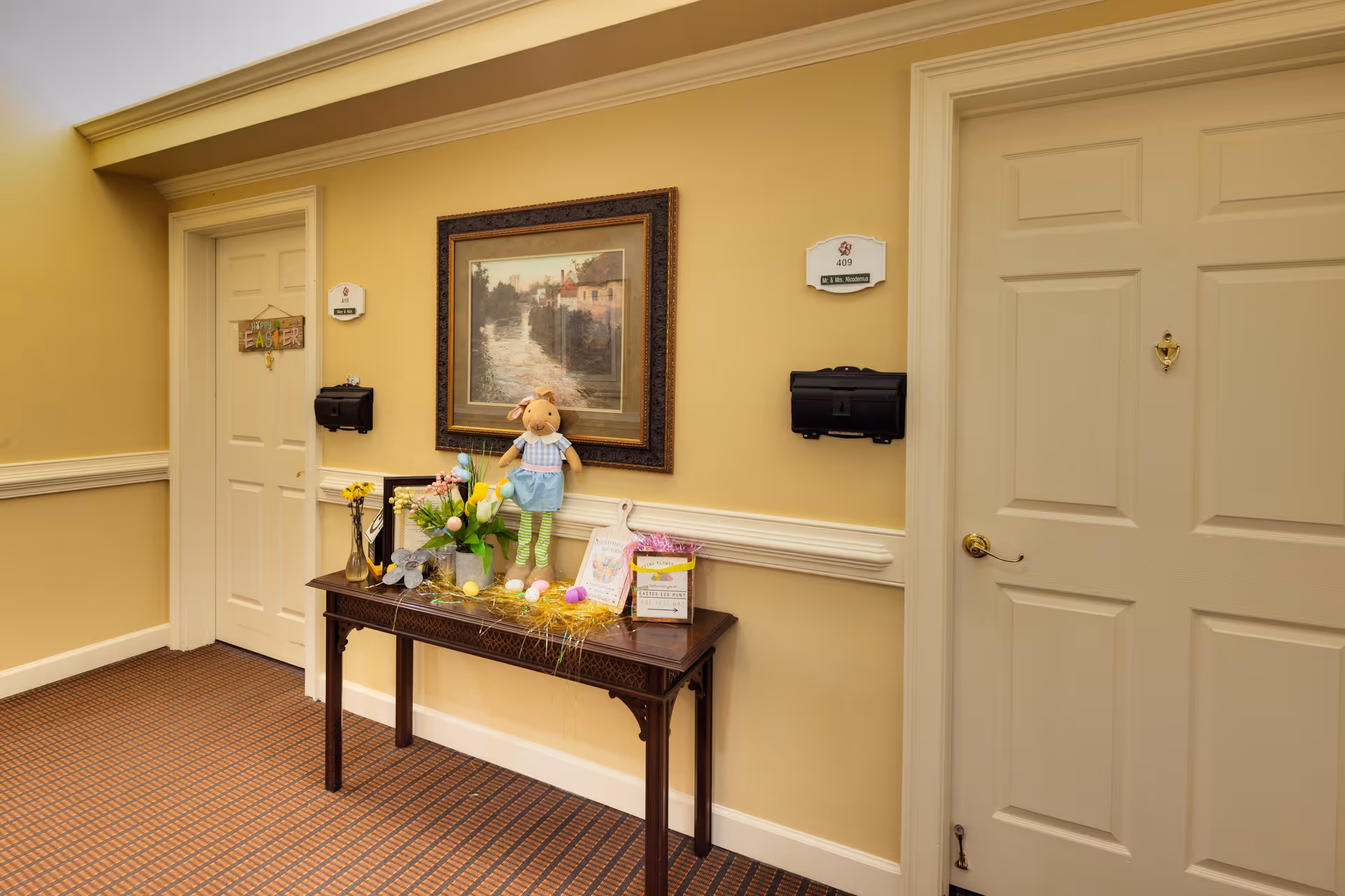 A hallway in a senior living facility with two closed doors, one decorated with an Easter sign. Between the doors is a wooden table adorned with Easter decorations including flowers, a doll, and colorful eggs. Above the table hangs a framed painting of a riverside scene. The walls are painted yellow with white trim, and the floor is carpeted in a patterned design.