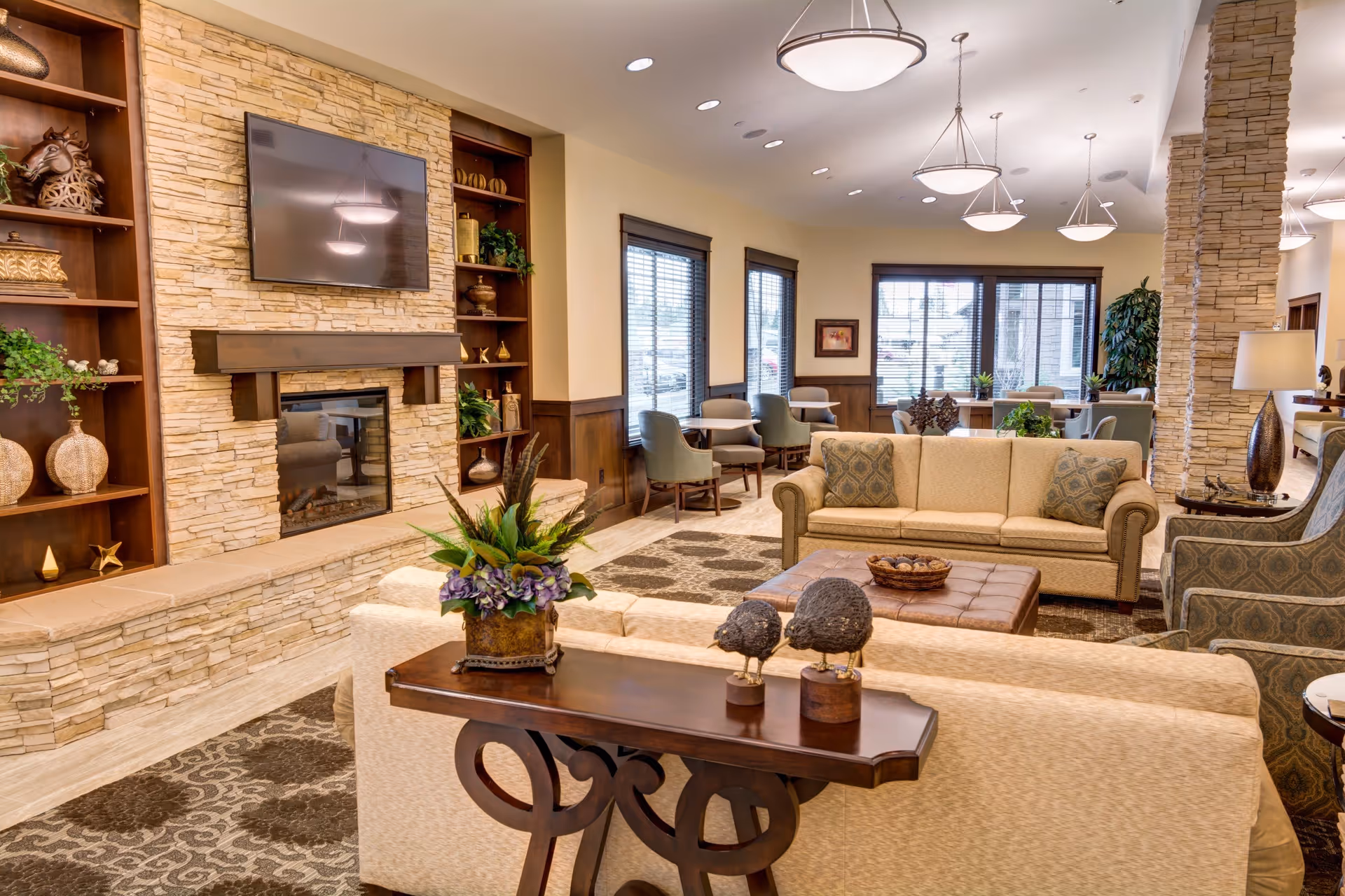 A spacious and well-lit living room area in an assisted living facility featuring beige sofas with patterned cushions, a wooden coffee table with decorative items, a stone fireplace with a mounted flat-screen TV above it, built-in wooden shelves with various decorative objects, large windows with blinds, and multiple ceiling light fixtures.