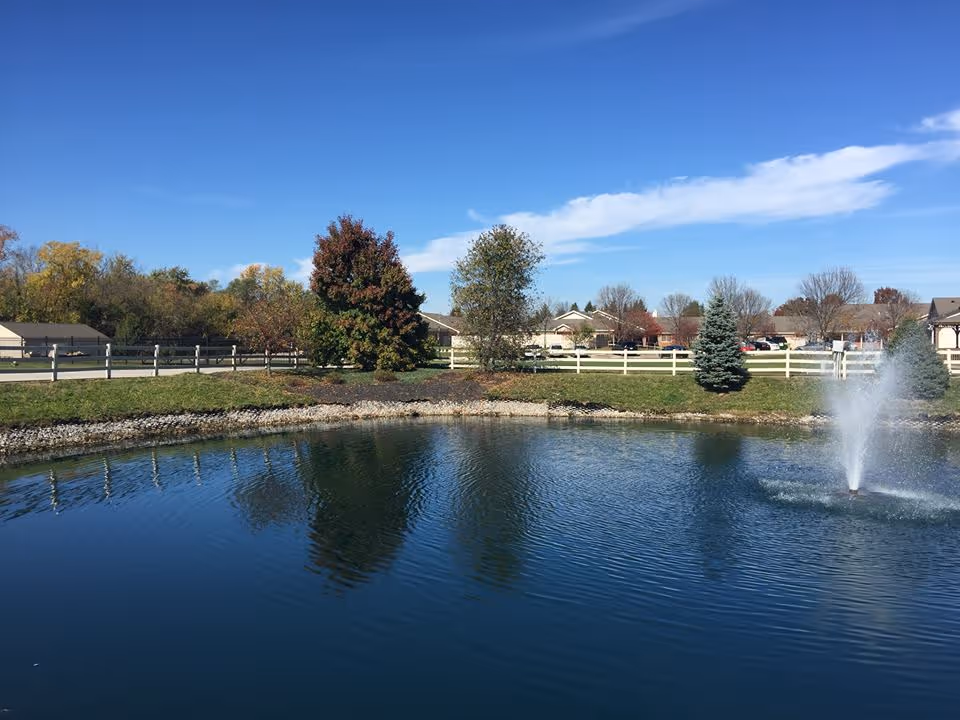 A serene outdoor scene featuring a small pond with a water fountain spraying water upwards. Surrounding the pond are green grassy areas, trees with autumn foliage, a white fence, and residential houses in the background under a clear blue sky with some clouds.