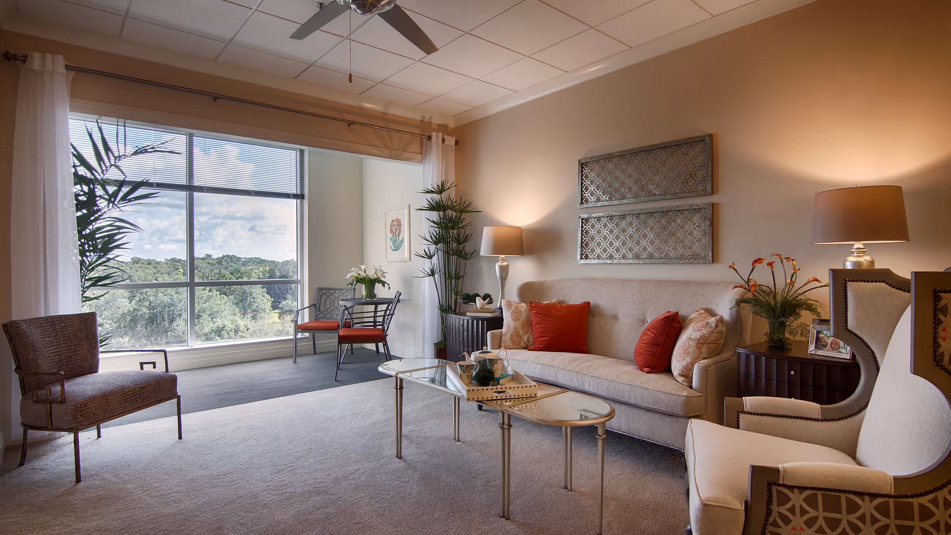 Sunlit, well-furnished living room with a sofa, armchairs, glass coffee table, lamps, plants, and a large window overlooking trees.