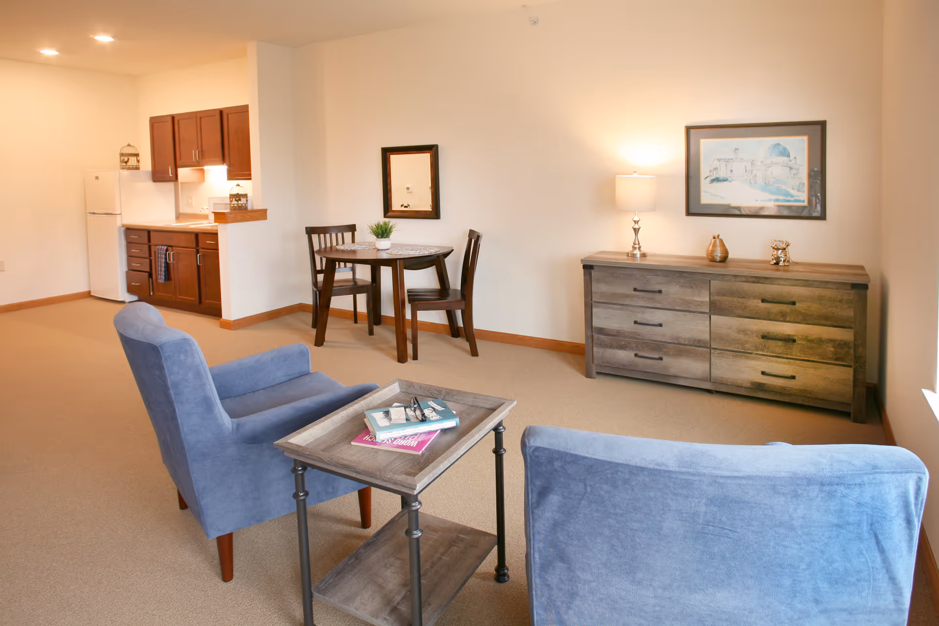 Cozy living area with two blue armchairs, a small coffee table, a dining set, kitchenette, and a dresser with a lamp and wall art.