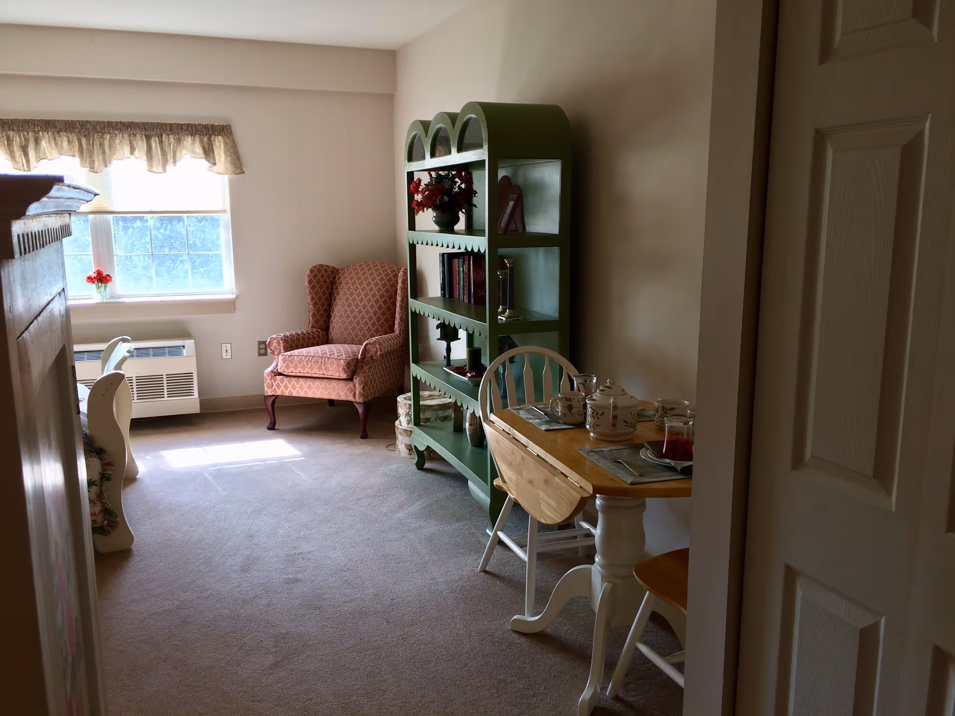 A cozy interior room with a window letting in natural light. There is a patterned armchair near the window, a green bookshelf filled with books and decorative items, and a small wooden dining table set with a teapot, cups, and placemats. The room has beige walls and carpeted floor.