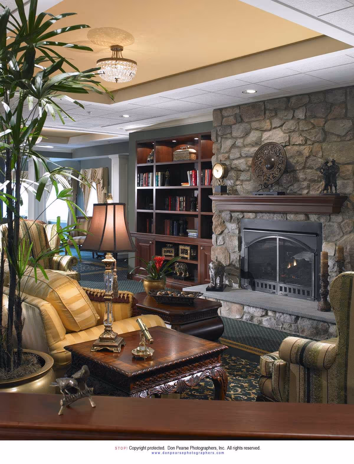 A cozy living room area with a stone fireplace, wooden mantel decorated with sculptures and a clock, a built-in bookshelf filled with books and decorative items, a yellow striped sofa, a wooden coffee table with a lamp and a small telescope, a potted plant, and an armchair with striped upholstery.