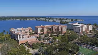 Aerial view of Westminster Shores senior living facility showing multiple buildings surrounded by greenery, a parking lot, and water in the background under a clear blue sky.