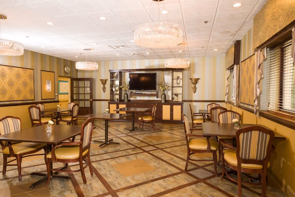 A well-lit dining room with several wooden tables and chairs upholstered in striped fabric. The room features patterned wallpaper with yellow and beige tones, three crystal chandeliers hanging from a white textured ceiling, and a large flat-screen TV mounted on a wooden cabinet with decorative vases and wall sconces on either side. Windows with blinds and valances are on the right side of the room.