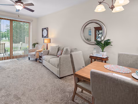 A cozy living and dining area in a senior living facility with a beige sofa, an armchair, a wooden dining table with four chairs, a round mirror on the wall, a small side table with a plant and decorative items, and large sliding glass doors with vertical blinds leading to an outdoor patio.